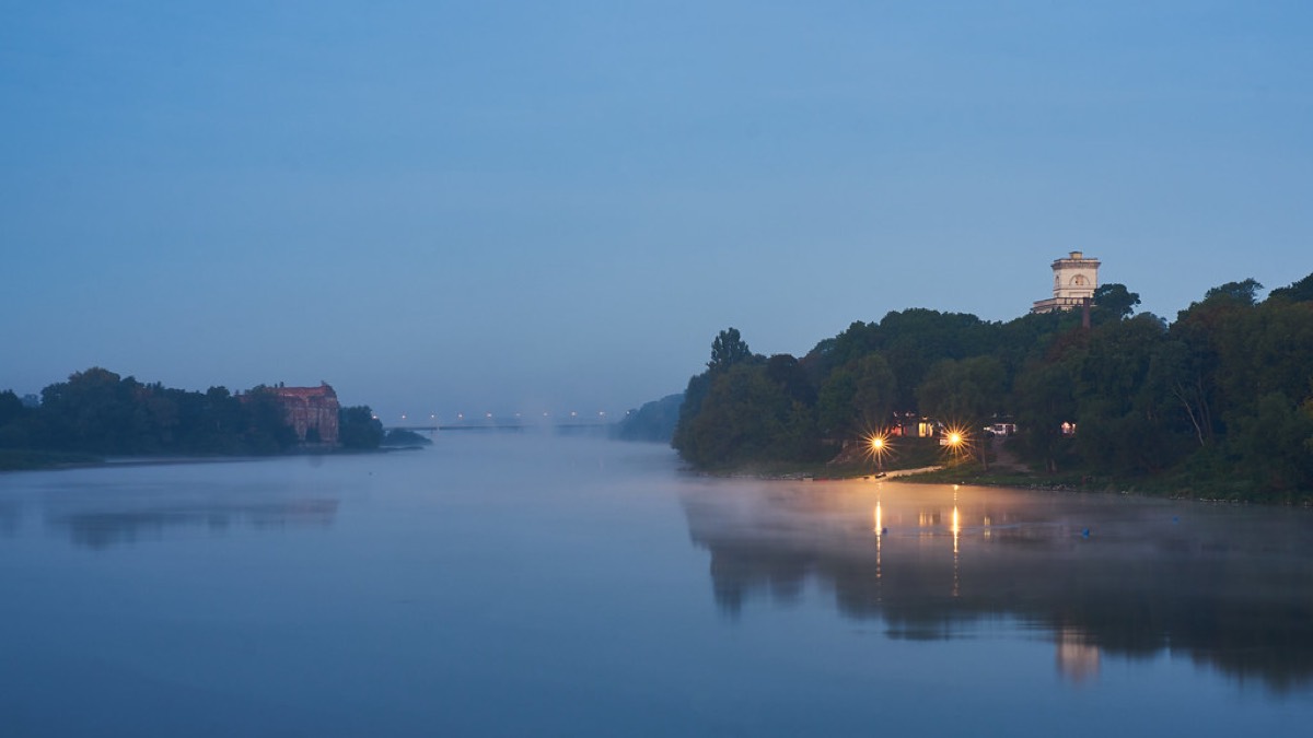 A calm river at first light, soft blue sky and mist rising off still water, with warm lamplight glowing on the far bank.
